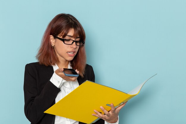 Front view female office worker in strict suit holding yellow document on light blue surface