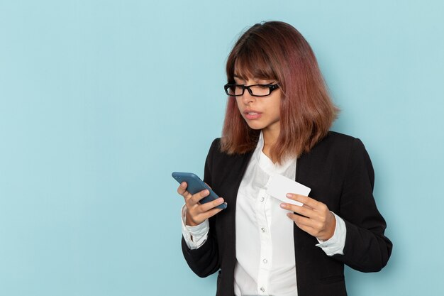 Front view female office worker using her phone and holding card on light-blue surface