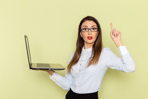 Front view of female office worker in white shirt and black skirt holding laptop on light-green wall