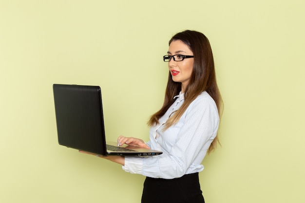 Front view of female office worker in white shirt and black skirt holding laptop on light green wall