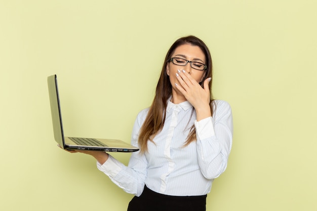 Front view of female office worker in white shirt and black skirt holding laptop and yawning on light-green wall