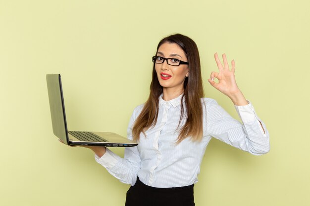 Front view of female office worker in white shirt and black skirt holding and using her laptop with smile on green wall