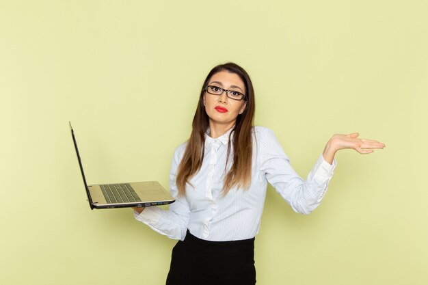 Front view of female office worker in white shirt and black skirt holding and using laptop on light-green wall