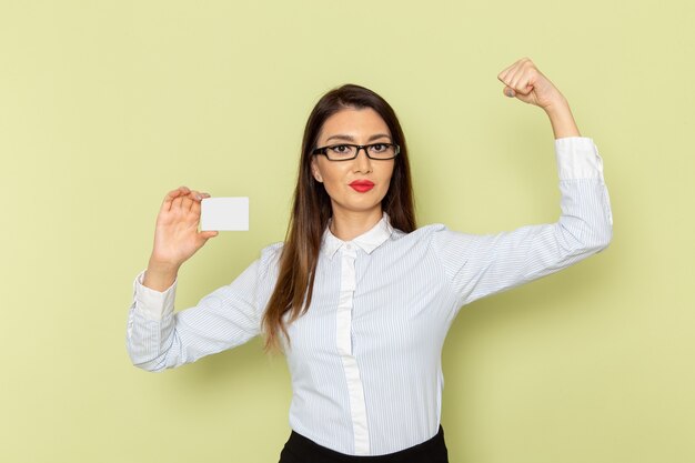 Front view of female office worker in white shirt and black skirt holding white card and flexing on light-green wall