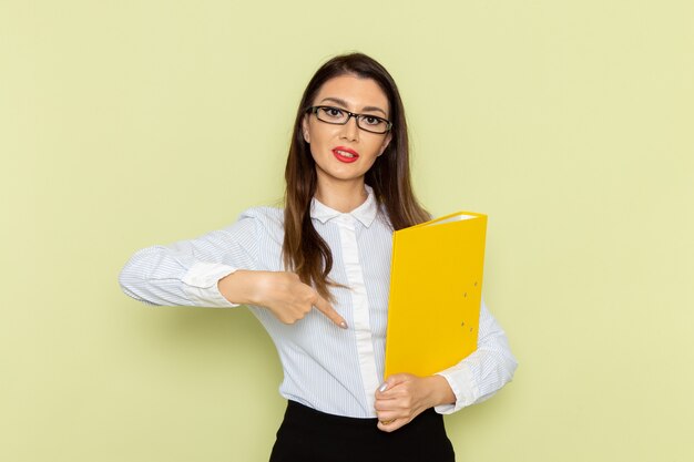 Front view of female office worker in white shirt and black skirt holding yellow file on green wall