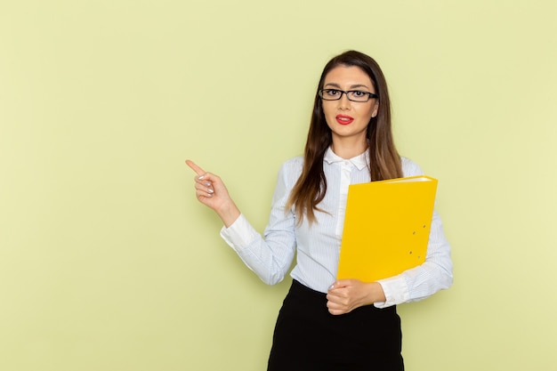 Front view of female office worker in white shirt and black skirt holding yellow file on green wall