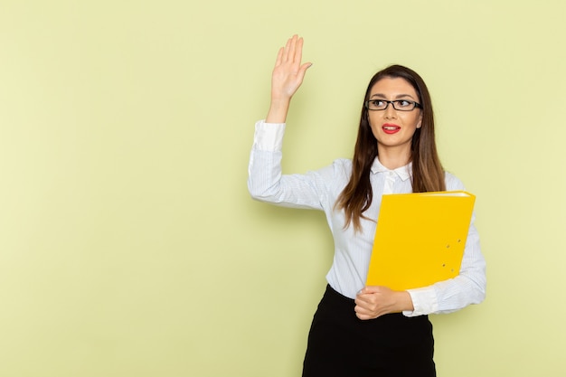 Front view of female office worker in white shirt and black skirt holding yellow file on light green wall