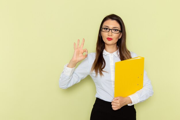 Front view of female office worker in white shirt and black skirt holding yellow file and posing on green wall