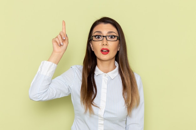 Front view of female office worker in white shirt and black skirt posing with raised finger on green wall