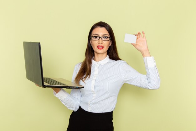 Front view of female office worker in white shirt and black skirt using her laptop and holding card on light-green wall