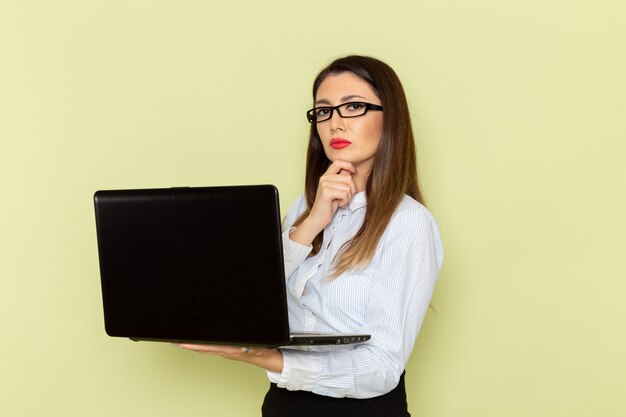 Front view of female office worker in white shirt and black skirt using her laptop on light-green wall