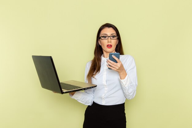 Front view of female office worker in white shirt and black skirt using her laptop on light green wall