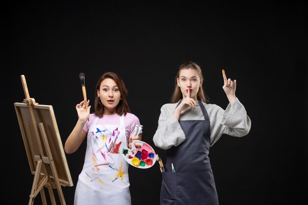 Vista frontale pittrice che disegna su cavalletto con un'altra donna sul muro nero foto a colori immagine artistica artista lavoro di pittura