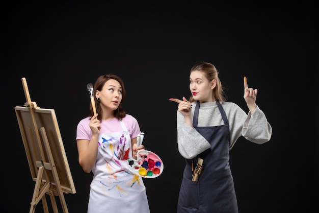 Free photo front view female painter drawing on easel with other female on black wall photo color art picture artist paints job draw