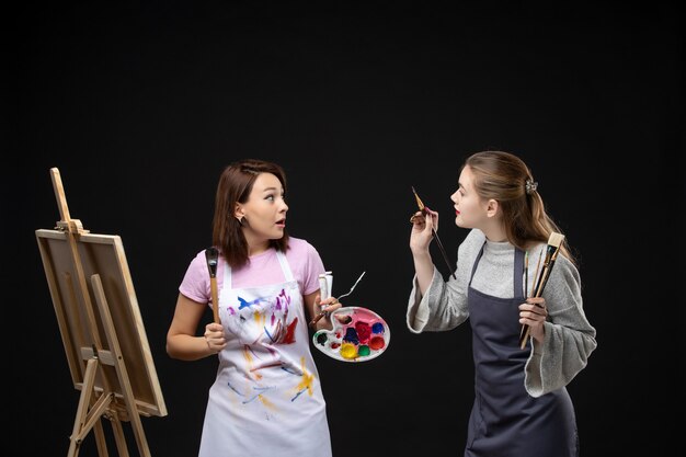 Foto gratuita vista frontale pittrici che tengono vernici e nappe per disegnare sulla parete nera pittura a colori disegnare lavoro immagine arte foto artista