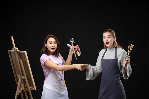 front view female painters holding paints and tassels for drawing shaking hands on black wall draw job picture art color photo painting artist