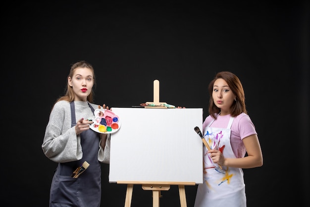 Front view of female painters with easel holding paints on dark wall