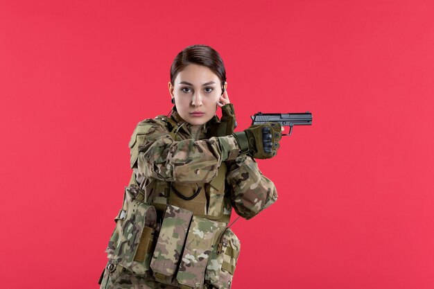 Вид спереди женщины-солдата в камуфляже, держащего пистолет на красной стене