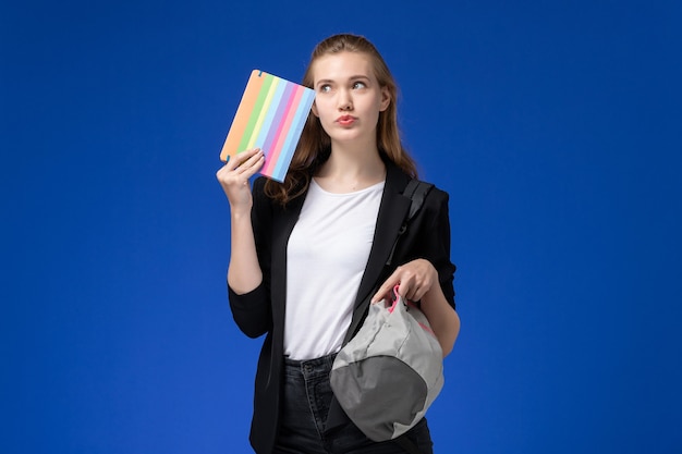 Front view female student in black jacket holding grey backpack and copybook on the blue wall school college university lesson time