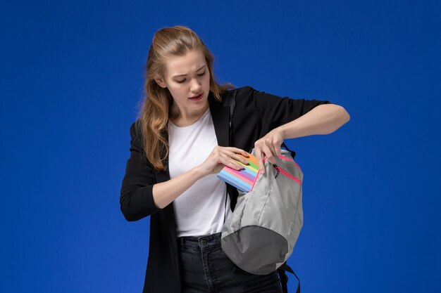 Front view female student in black jacket holding grey backpack and copybook putting it inside on the blue wall school college university lesson time