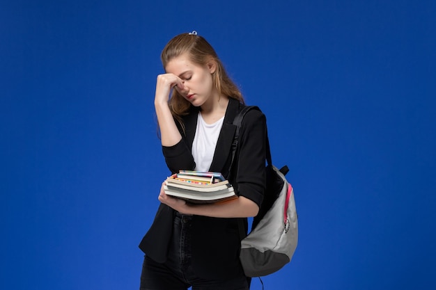 Front view female student in black jacket wearing backpack holding books on the blue wall school university college lessons