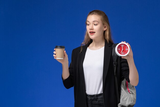 Front view female student in black jacket wearing backpack holding clocks and coffee on blue wall school college university lesson