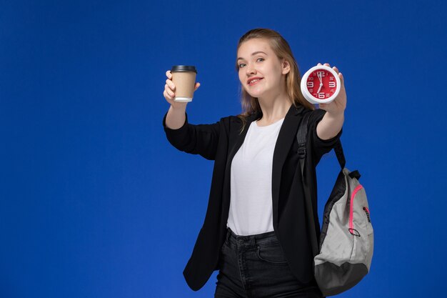 Front view female student in black jacket wearing backpack holding clocks and coffee smiling on blue wall school college university lesson