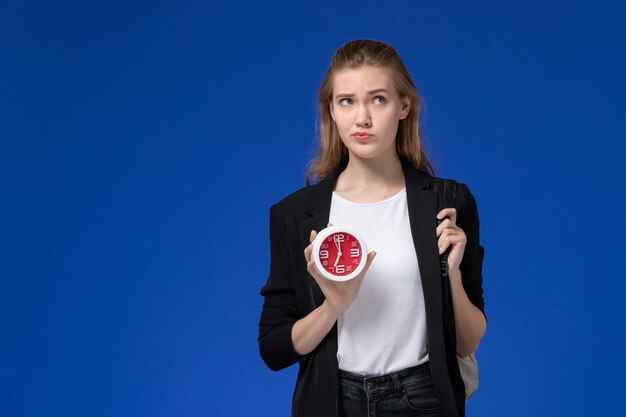 Front view female student in black jacket wearing backpack holding clocks thinking on blue wall school college university lesson