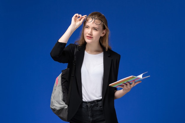 Free photo front view female student in black jacket wearing backpack holding copybook on the blue wall lesson books school college university