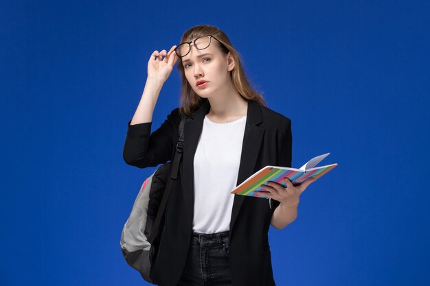 Front view female student in black jacket wearing backpack holding copybook on blue wall lesson school college university