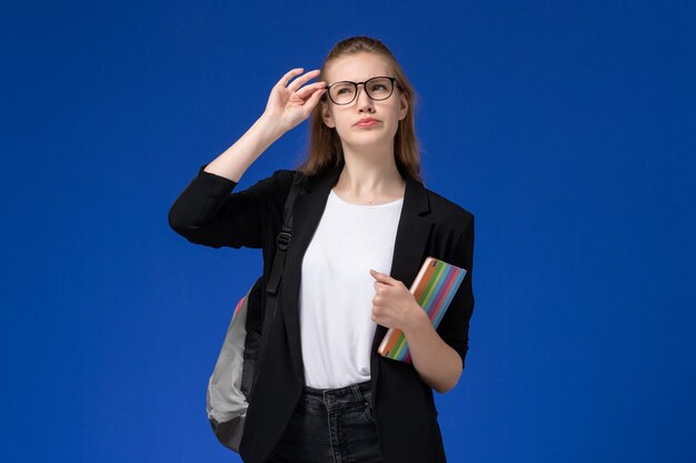 Front view female student in black jacket wearing backpack holding copybook on blue wall lessons school college university