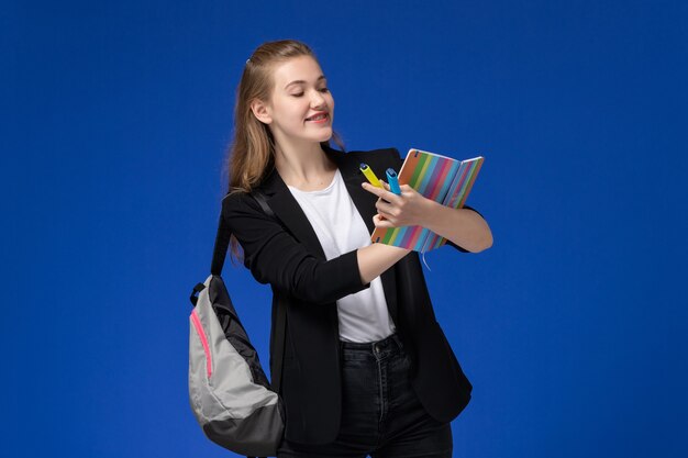 Front view female student in black jacket wearing backpack holding felt pens and copybook on the blue wall lessons school college university