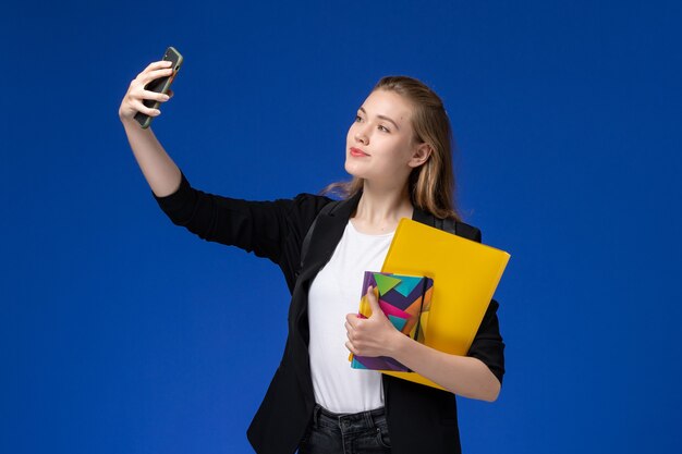 Front view female student in black jacket wearing backpack holding file and copybook taking selfie on blue wall college university lessons