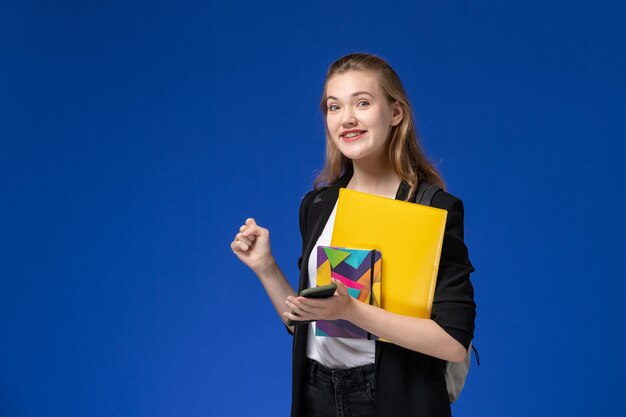 青い壁の大学のレッスンでファイルと電話でコピーブックを保持しているバックパックを身に着けている黒いジャケットの正面図女子学生