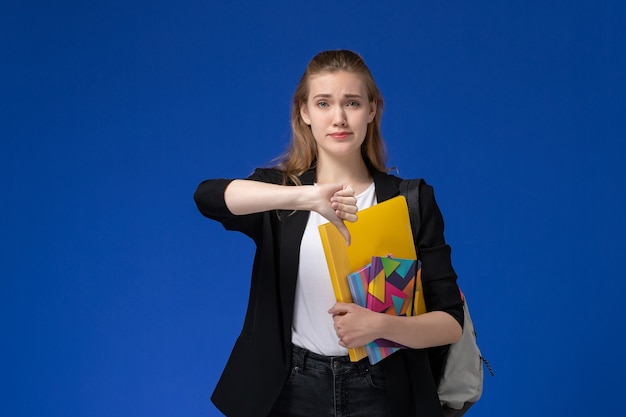 Front view female student in black jacket wearing backpack holding files with copybooks on the blue wall college university lessons
