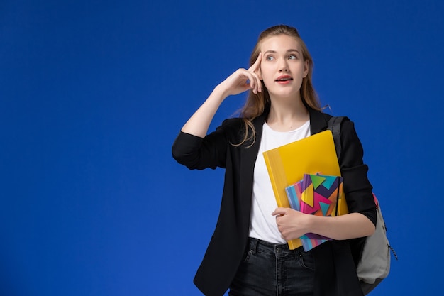 Studentessa di vista frontale in giacca nera che indossa lo zaino che tiene i file con i quaderni pensando sulle lezioni dell'università universitaria della parete blu