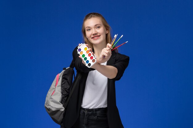 Front view female student in black jacket wearing backpack holding paints for drawing and tassel on blue wall drawing art school college lessons