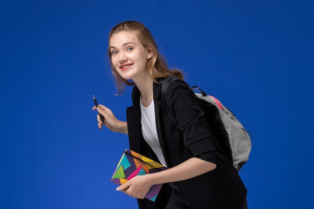 Вид спереди студентка в черной куртке, носить рюкзак, держа ручку и тетрадь, улыбаясь на синей стене книги уроки школы колледжа университета