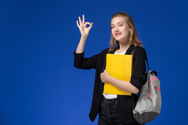 Front view female student in black jacket wearing backpack and holding yellow file on blue wall college university lesson