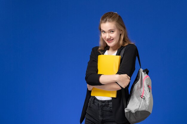 Front view female student in black jacket wearing backpack holding yellow file on blue wall school college university lesson