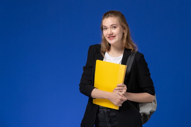 Front view female student in black jacket wearing backpack and holding yellow file on the blue wall school college university lessons