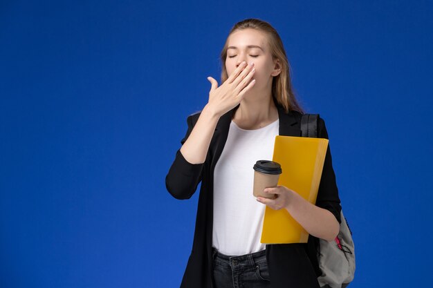 Front view female student in black jacket wearing backpack holding yellow file and coffee yawning on blue wall school college university lesson