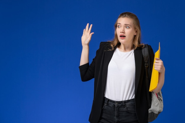 Вид спереди студентка в черной куртке и рюкзаке с желтыми папками на голубой стене, школьный колледж, университетский урок