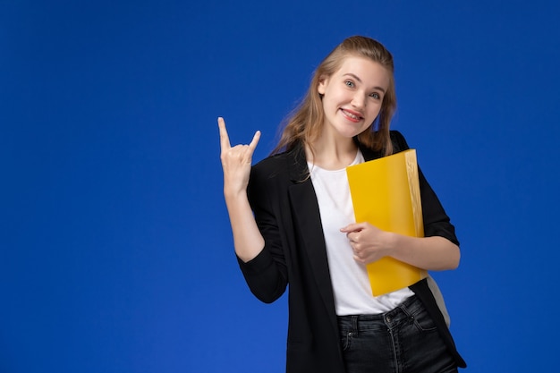 Front view female student in black jacket wearing backpack holding yellow files and smiling on blue wall school college university lesson