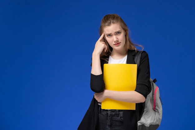 Front view female student in black jacket wearing backpack holding yellow files thinking on blue wall school college university lesson