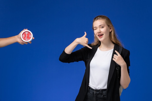 Front view female student in black jacket wearing backpack posing on the blue wall school college university lessons time