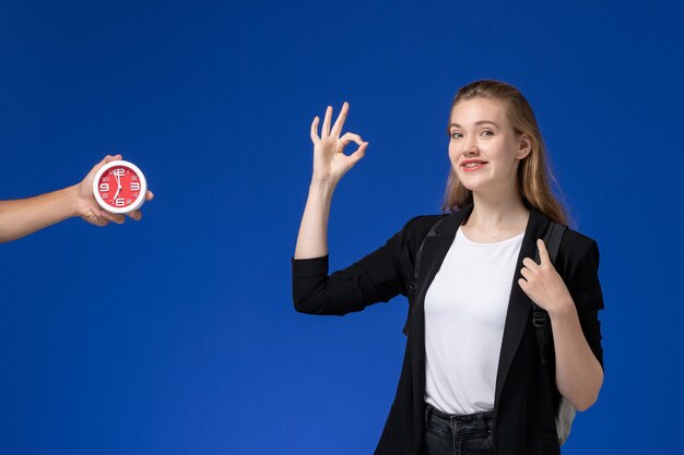 Front view female student in black jacket wearing backpack showing alright sign on blue wall school college university lessons time