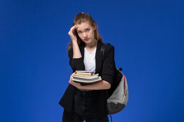 Front view female student in black jacket wearing backpack thinking and holding books on the blue wall drawing art school college