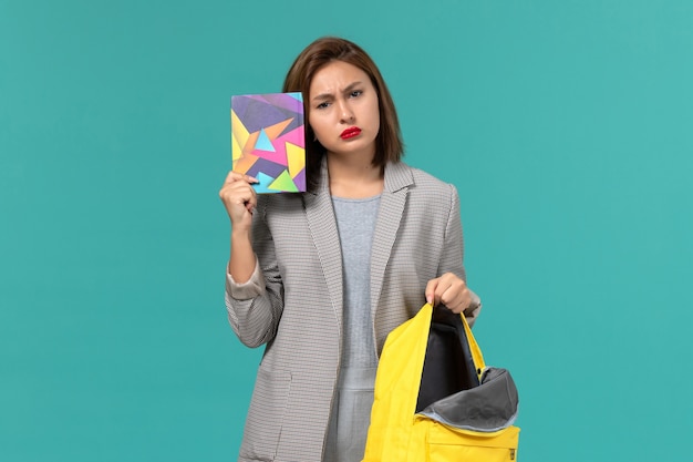 Front view of female student in grey jacket holding yellow backpack and copybook on the light blue wall