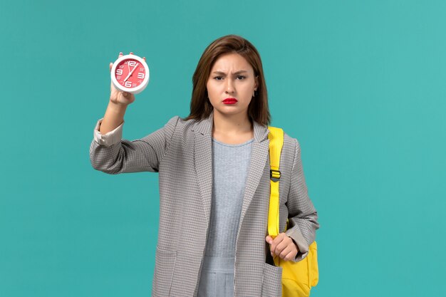 Free photo front view of female student in grey jacket wearing her yellow backpack holding clocks on blue wall